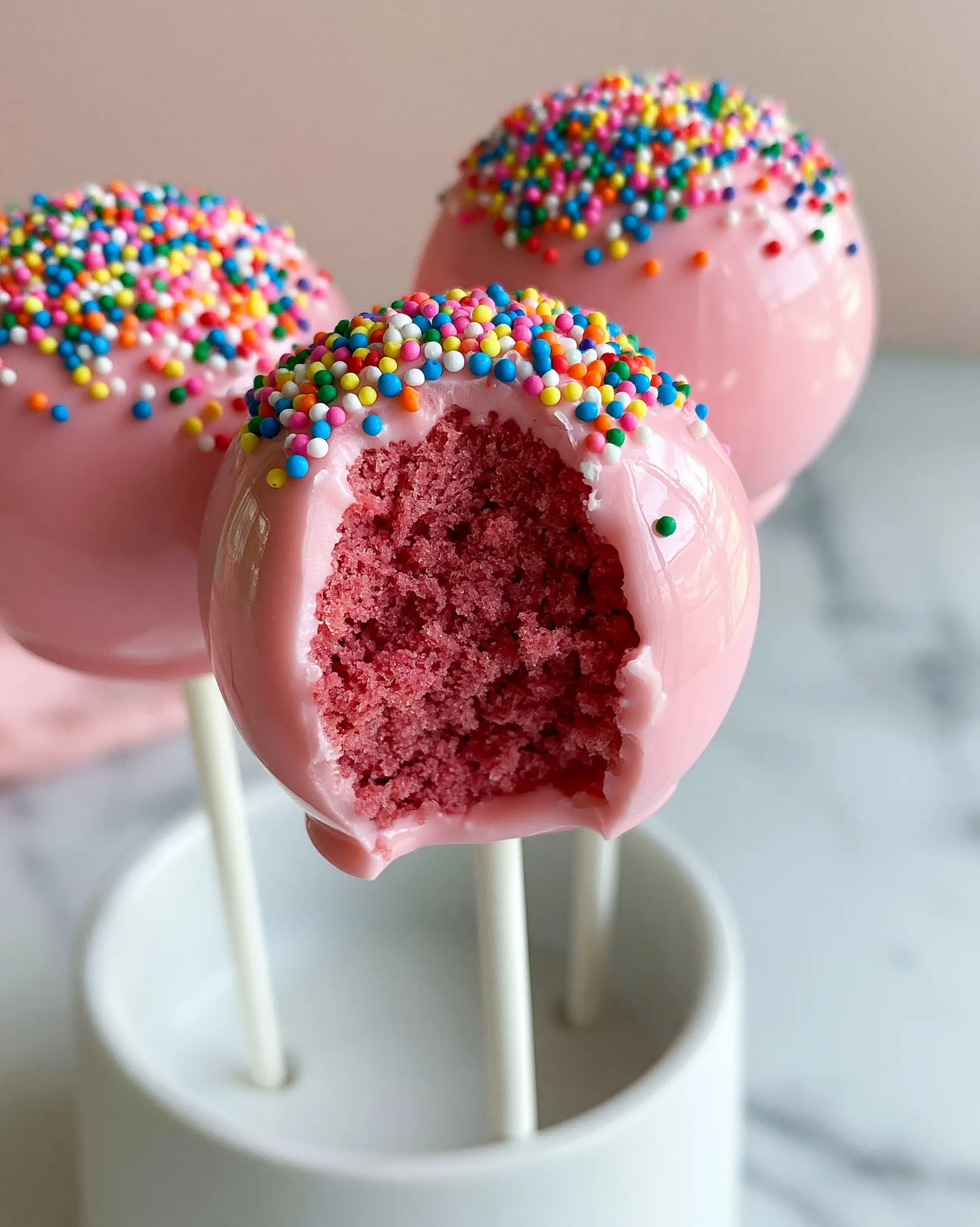 Three Birthday Cake Pops in a white minimalist stand, showing a smooth crack-free pastel pink candy coating and colorful nonpareil sprinkles.